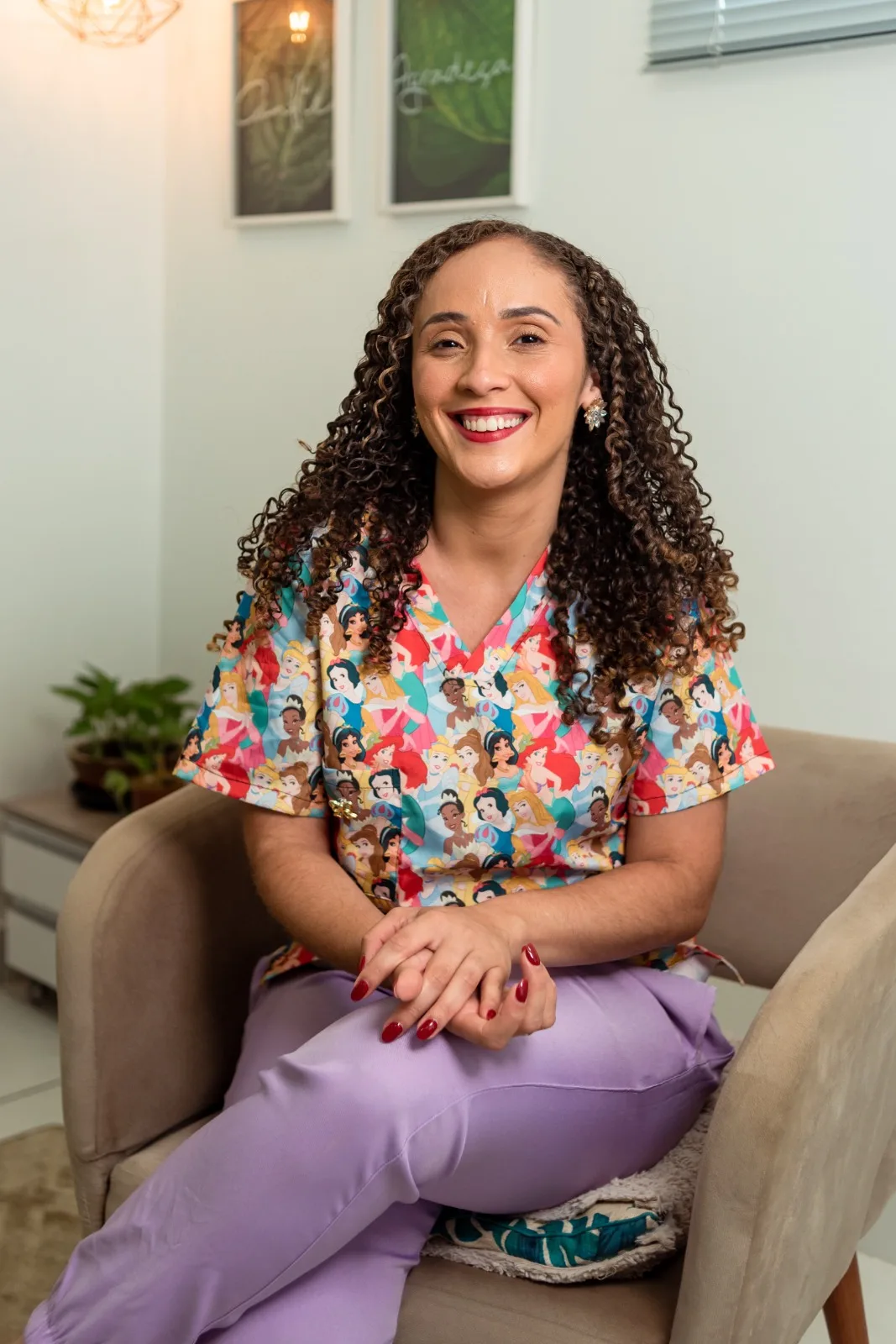 Milena Santos sentada em uma poltrona bege, sorrindo para a câmera, com blusa colorida estampada e calça lilás